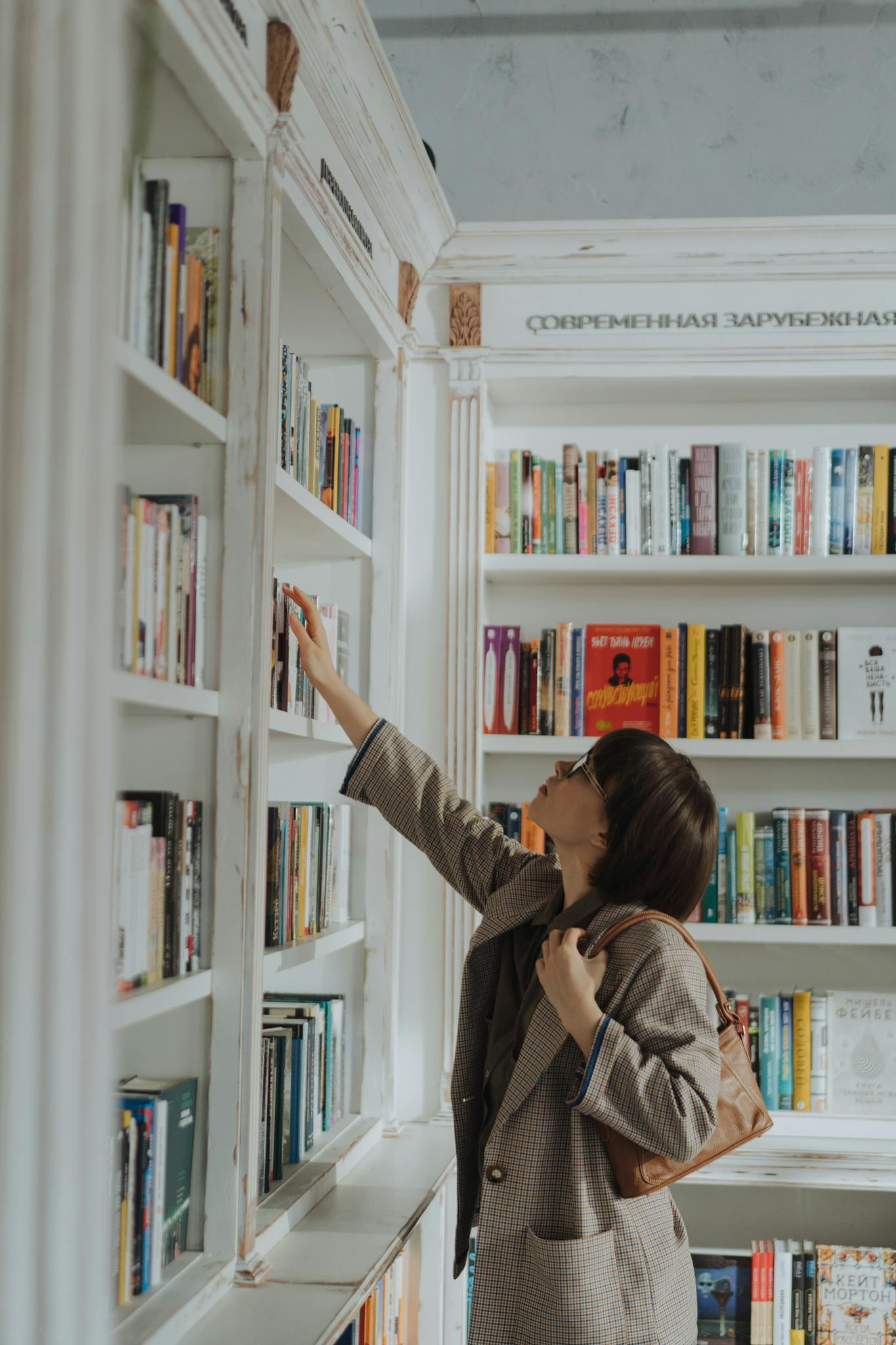 A young woman reaching for a book on a stylish library bookshelf, embodying education and leisure.